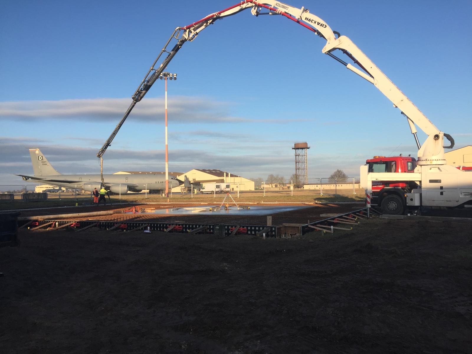 Concrete pour using a boom pump at RAF Mildenhall, with a US Air Force aircraft and airfield buildings beyond