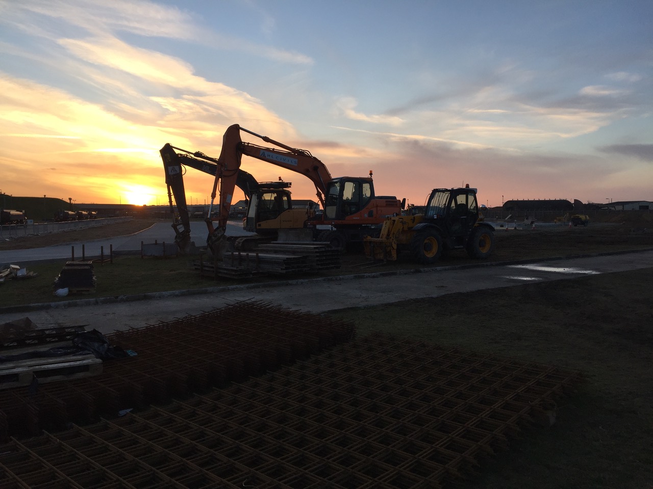 Excavators and a telehandler working at sunset on the RAF Lakenheath refurbishment, with stacked reinforcement mesh in the foreground