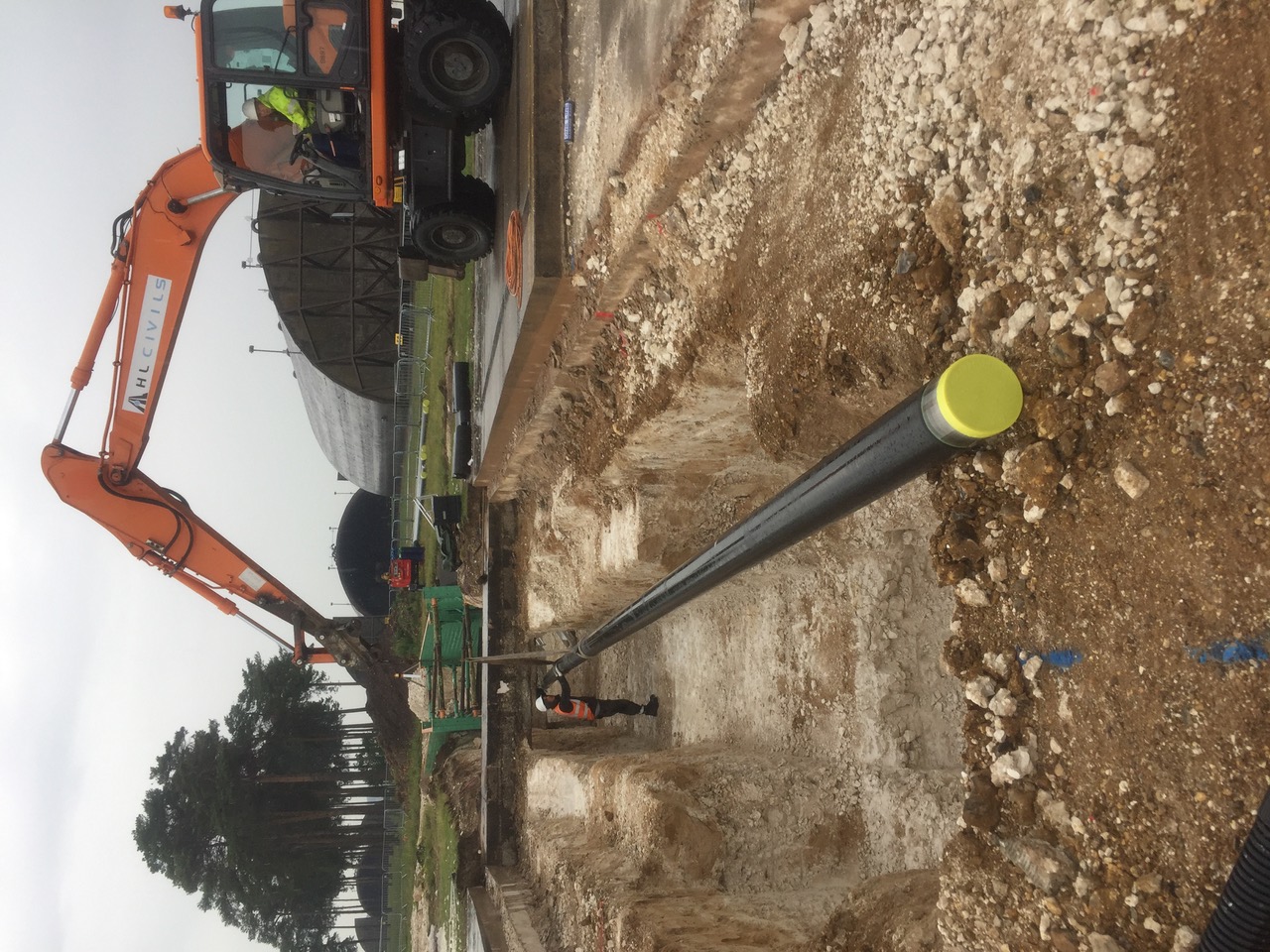 HL Civils excavator lowering pipework into an open fuel-main trench at RAF Lakenheath